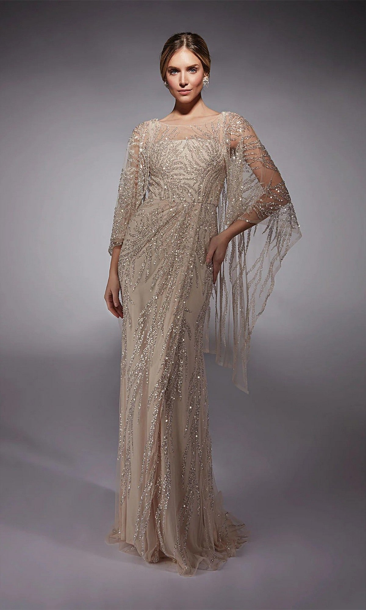 A woman models the Alyce 27754, a floor-length pink champagne formal gown with intricate beading and sheer draped sleeves—an elegant choice for a distinguished mother-of-the-bride.